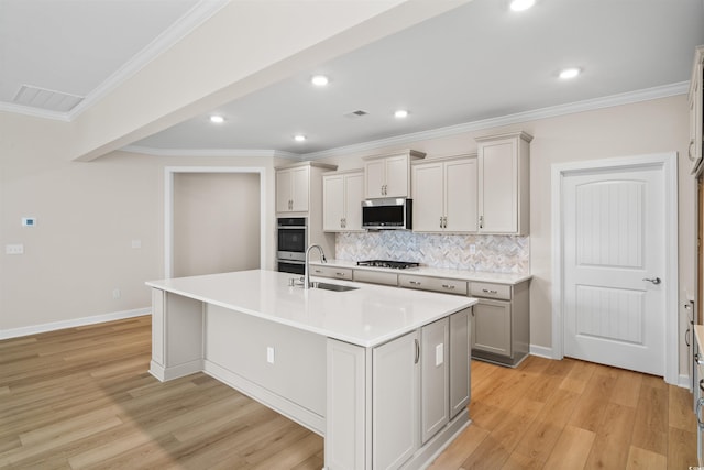 kitchen featuring appliances with stainless steel finishes, a sink, light wood finished floors, an island with sink, and tasteful backsplash
