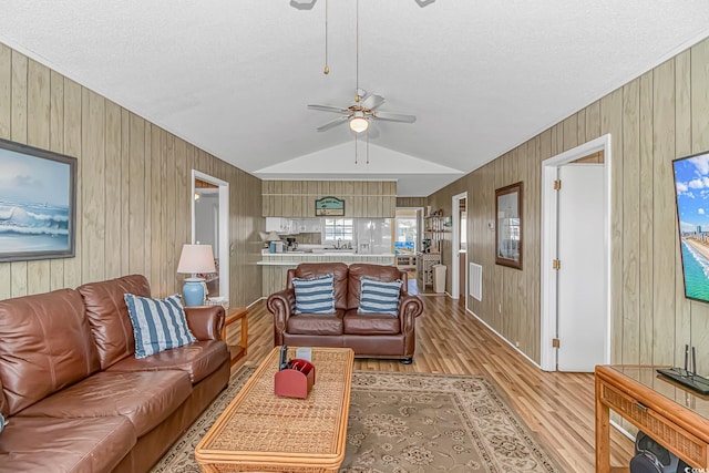 living area with light wood-style flooring, a ceiling fan, lofted ceiling, wooden walls, and a textured ceiling