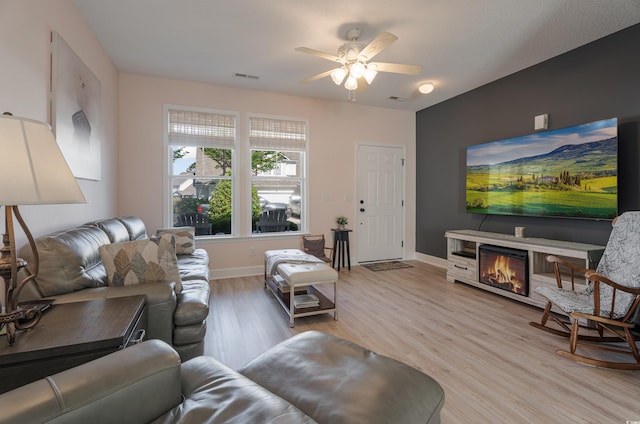 living room featuring a ceiling fan, light wood-type flooring, baseboards, and a glass covered fireplace