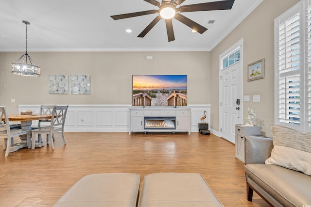 living room featuring crown molding, light wood-style flooring, ceiling fan, a chandelier, and a glass covered fireplace