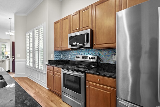 kitchen with appliances with stainless steel finishes, ornamental molding, dark stone countertops, a wainscoted wall, and brown cabinetry