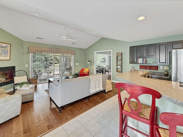 living area with vaulted ceiling, a textured ceiling, a glass covered fireplace, light tile patterned floors, and ceiling fan
