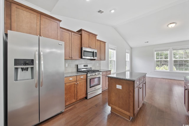 kitchen with appliances with stainless steel finishes, vaulted ceiling, brown cabinets, light stone counters, and dark wood finished floors