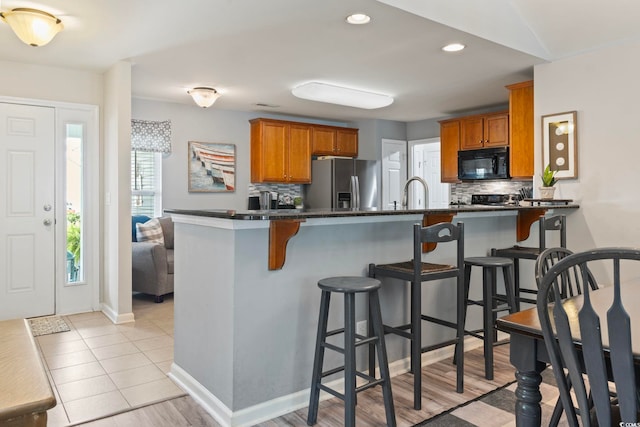 kitchen with black microwave, stainless steel fridge with ice dispenser, dark countertops, a kitchen bar, and recessed lighting