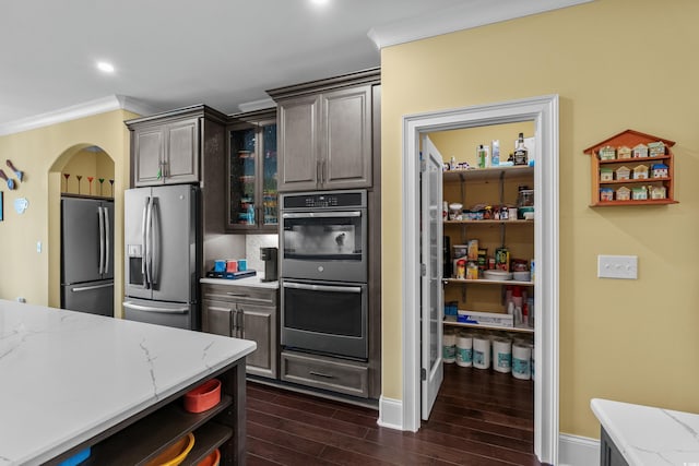 kitchen with appliances with stainless steel finishes, dark wood-style floors, ornamental molding, light stone countertops, and glass insert cabinets