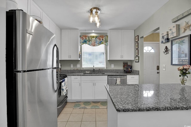 kitchen with appliances with stainless steel finishes, a sink, white cabinetry, light tile patterned floors, and dark stone countertops