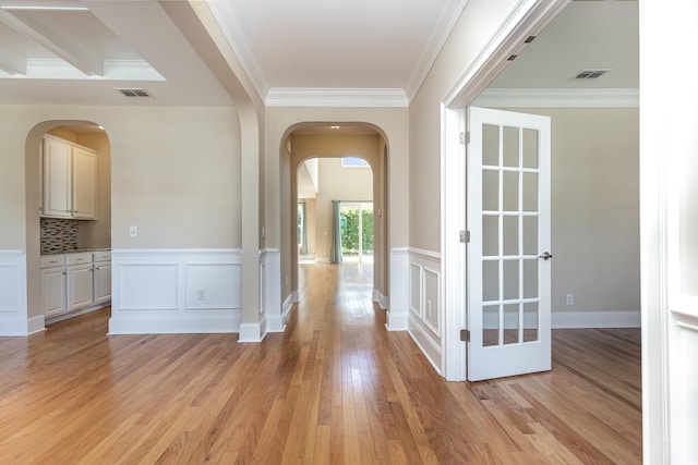 hallway with arched walkways, light wood-style floors, crown molding, wainscoting, and a decorative wall