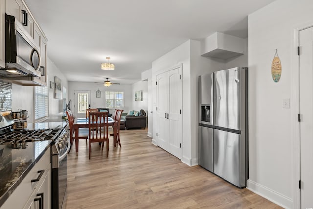kitchen with appliances with stainless steel finishes, a ceiling fan, light wood-style flooring, dark stone counters, and baseboards