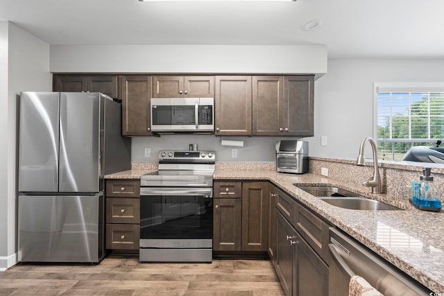 kitchen featuring appliances with stainless steel finishes, a sink, dark brown cabinets, light stone counters, and light wood-type flooring
