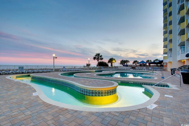 pool at dusk featuring a hot tub, a community pool, a patio area, and fence