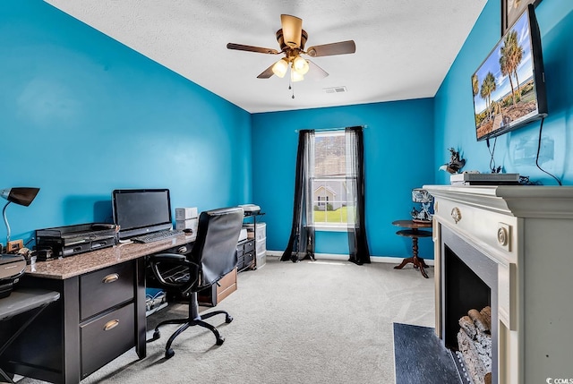 home office featuring light colored carpet, a fireplace with flush hearth, ceiling fan, baseboards, and a textured ceiling