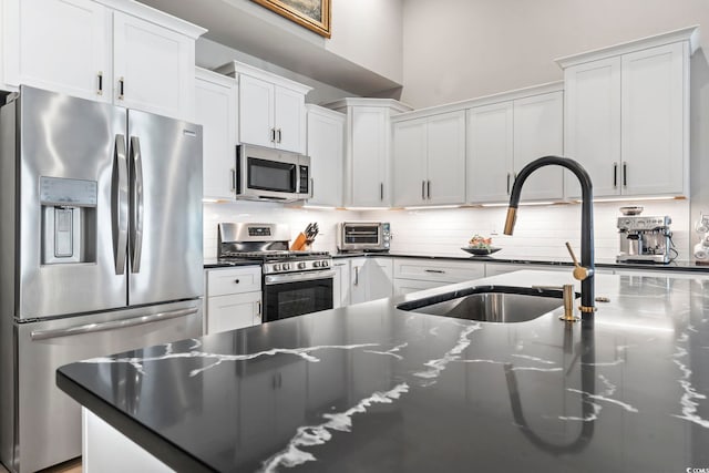 kitchen featuring appliances with stainless steel finishes, a sink, backsplash, white cabinets, and dark stone counters
