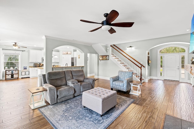 living room featuring arched walkways, ceiling fan, stairs, wood finished floors, and ornamental molding