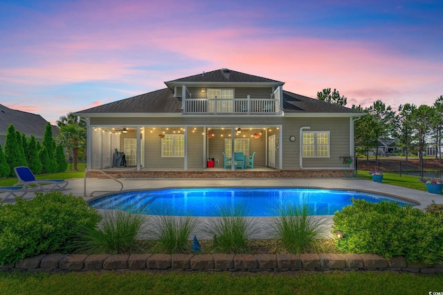 back of property at dusk featuring a ceiling fan, a balcony, an outdoor pool, and a patio