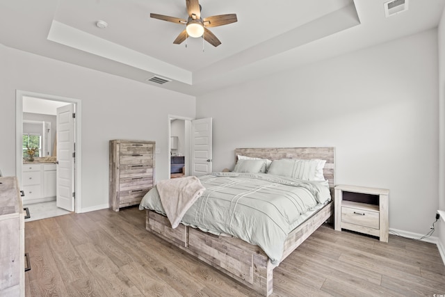 bedroom featuring a tray ceiling, wood finished floors, baseboards, ensuite bathroom, and ceiling fan