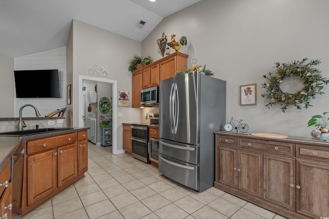 kitchen with appliances with stainless steel finishes, a sink, light tile patterned flooring, brown cabinetry, and high vaulted ceiling