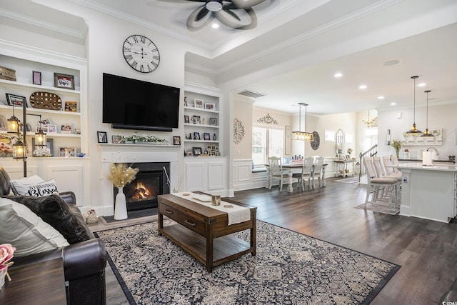 living room featuring crown molding, a ceiling fan, dark wood-style floors, a fireplace with flush hearth, and built in shelves