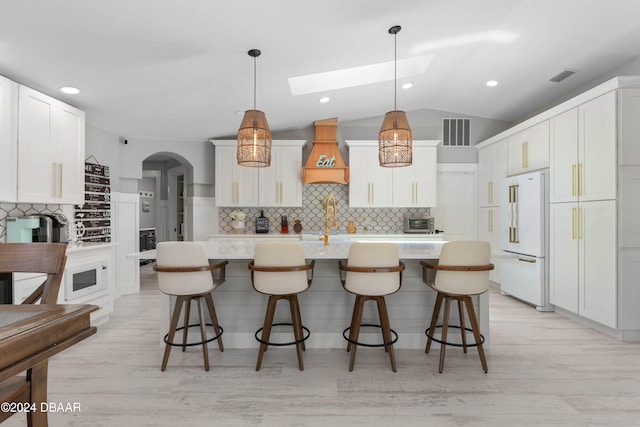 kitchen with light wood-type flooring, custom exhaust hood, high end white fridge, a center island with sink, and hanging light fixtures