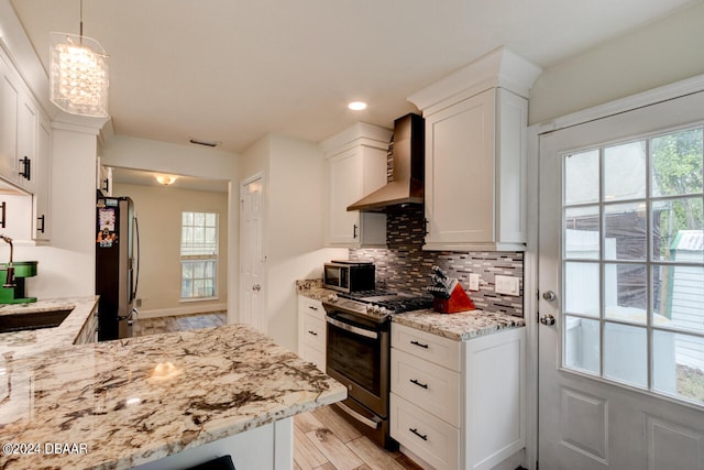 kitchen featuring stainless steel appliances, a healthy amount of sunlight, wall chimney range hood, pendant lighting, and white cabinetry