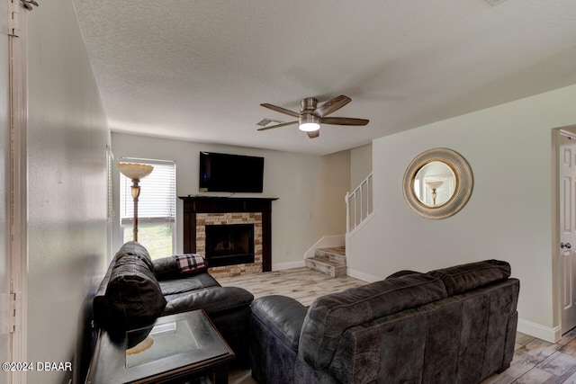 living room with ceiling fan, a stone fireplace, a textured ceiling, and light hardwood / wood-style flooring