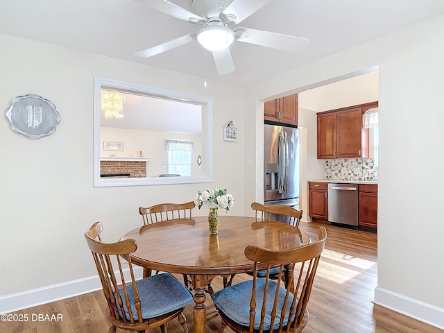 dining room featuring light wood-type flooring, a brick fireplace, baseboards, and a ceiling fan