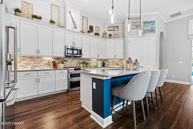 kitchen with hanging light fixtures, ornamental molding, appliances with stainless steel finishes, a kitchen island with sink, and white cabinets