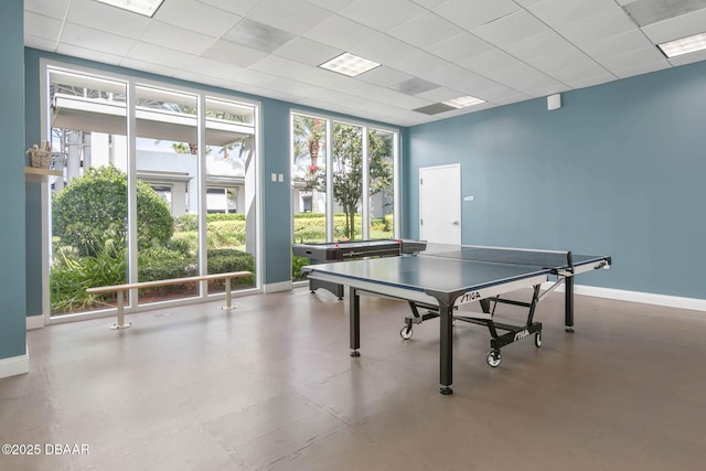 recreation room featuring a wall of windows, a drop ceiling, finished concrete flooring, and baseboards