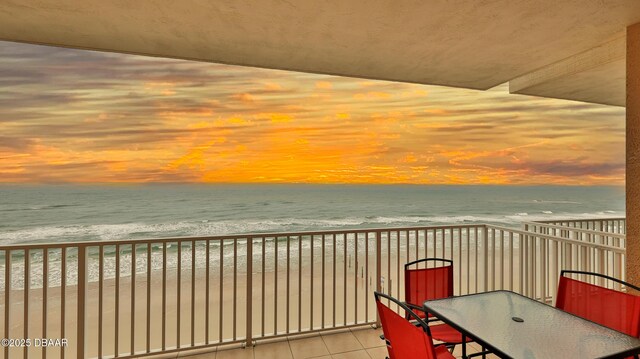 balcony at dusk featuring outdoor dining area, a water view, and a view of the beach