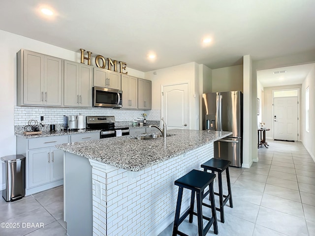 kitchen with light tile patterned floors, sink, dark stone countertops, stainless steel appliances, and a center island with sink