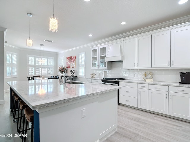 kitchen featuring ornamental molding, white cabinetry, custom range hood, hanging light fixtures, and an island with sink