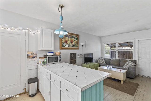 kitchen with tile counters, white cabinets, stainless steel microwave, hanging light fixtures, and a peninsula