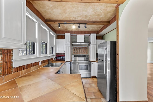 kitchen with arched walkways, appliances with stainless steel finishes, wall chimney range hood, white cabinetry, and a sink