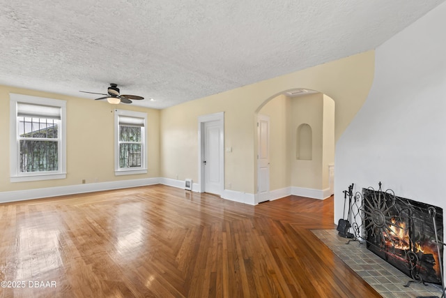 unfurnished living room featuring arched walkways, ceiling fan, a textured ceiling, a fireplace with flush hearth, and baseboards