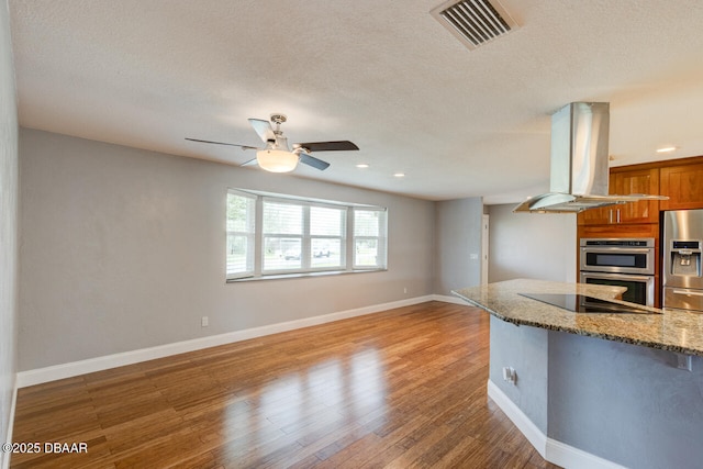 kitchen with a textured ceiling, hardwood / wood-style flooring, island exhaust hood, stainless steel appliances, and light stone countertops