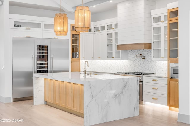 kitchen featuring high end appliances, light wood-style flooring, backsplash, a kitchen island with sink, and light stone countertops