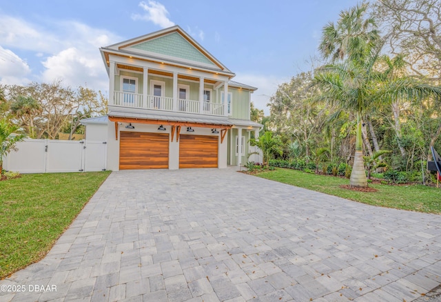 coastal inspired home with an attached garage, a balcony, decorative driveway, a gate, and a front yard