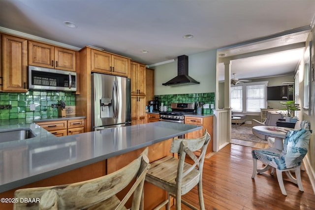 kitchen with brown cabinetry, stainless steel appliances, decorative backsplash, light wood-style floors, and wall chimney exhaust hood