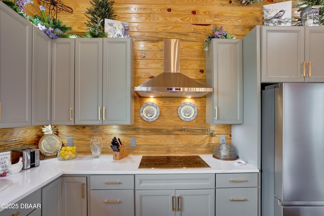 kitchen featuring gray cabinetry, black cooktop, stainless steel refrigerator, and wall chimney exhaust hood