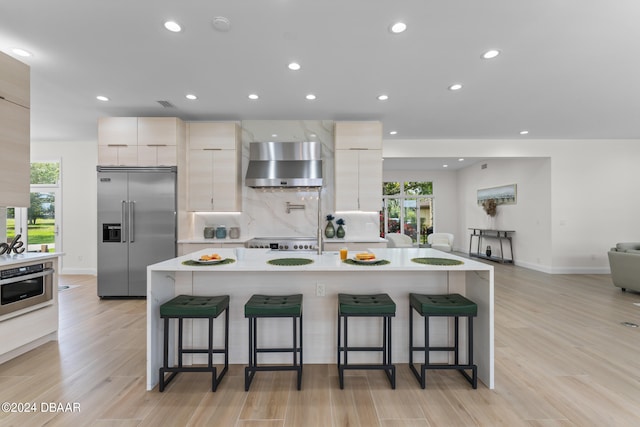 kitchen with appliances with stainless steel finishes, an island with sink, light hardwood / wood-style floors, a breakfast bar area, and wall chimney exhaust hood