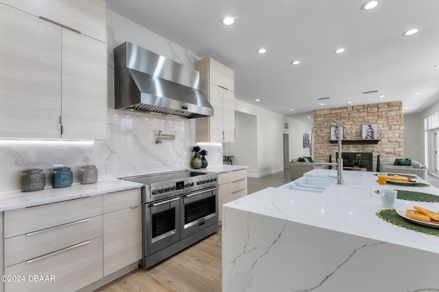 kitchen with light stone counters, a stone fireplace, range with two ovens, wall chimney range hood, and light wood-type flooring