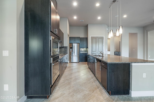 kitchen with a sink, appliances with stainless steel finishes, an island with sink, dark stone countertops, and decorative light fixtures