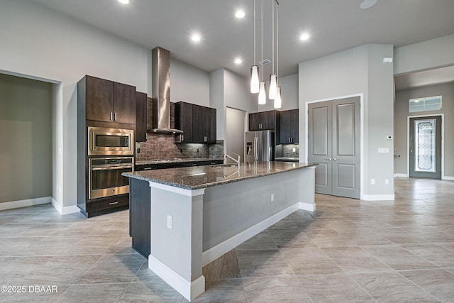 kitchen with decorative light fixtures, stainless steel appliances, a kitchen island with sink, dark brown cabinetry, and wall chimney range hood