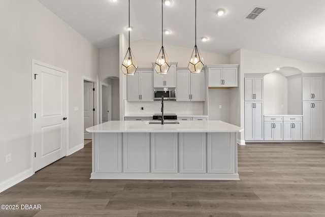 kitchen featuring stainless steel microwave, arched walkways, light countertops, an island with sink, and high vaulted ceiling