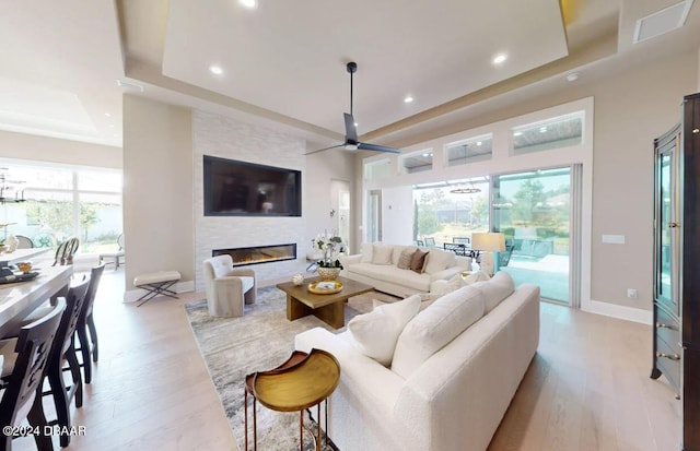 living room with a large fireplace, a tray ceiling, light hardwood / wood-style flooring, and ceiling fan