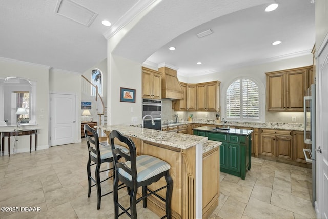 kitchen with sink, ornamental molding, a kitchen island with sink, light stone counters, and stainless steel double oven