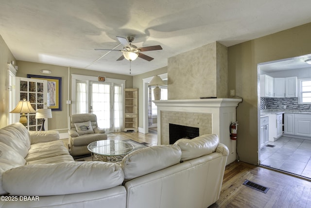 living area featuring ceiling fan, a brick fireplace, light wood-style flooring, baseboards, and visible vents