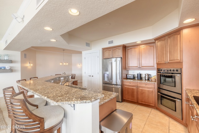 kitchen featuring stainless steel appliances, a center island with sink, a textured ceiling, light tile patterned flooring, and light stone countertops