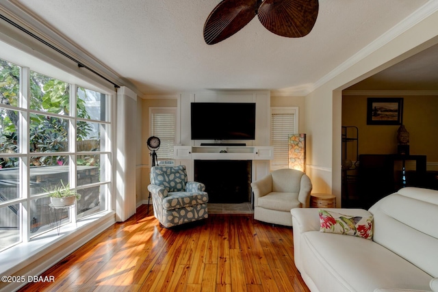 living area featuring a fireplace with raised hearth, ceiling fan, a textured ceiling, wood finished floors, and crown molding