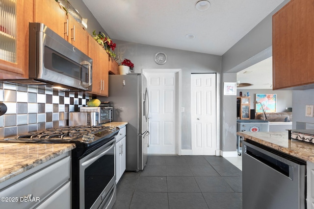 kitchen featuring brown cabinetry, appliances with stainless steel finishes, dark tile patterned floors, lofted ceiling, and backsplash