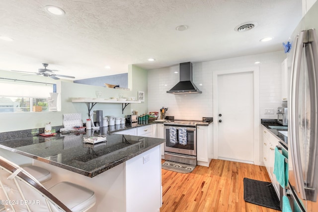 kitchen featuring stainless steel appliances, white cabinets, kitchen peninsula, wall chimney exhaust hood, and light hardwood / wood-style flooring